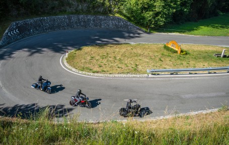 Three motorcycles are driving on a winding mountain road with guardrails and grassy areas.