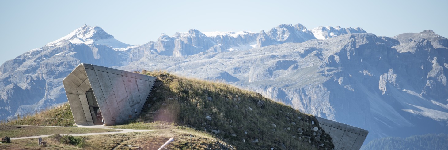 Moderno edificio in cemento inclinato su una collina erbosa davanti a montagne innevate sotto un cielo sereno.