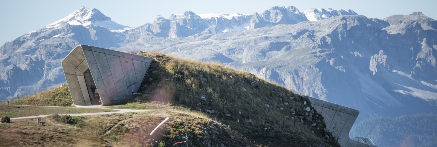 Modern, slanting concrete building on a grassy hill in front of snow-covered mountains under a clear sky.