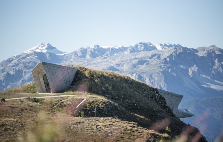 Modern, slanting concrete building on a grassy hill in front of snow-covered mountains under a clear sky.