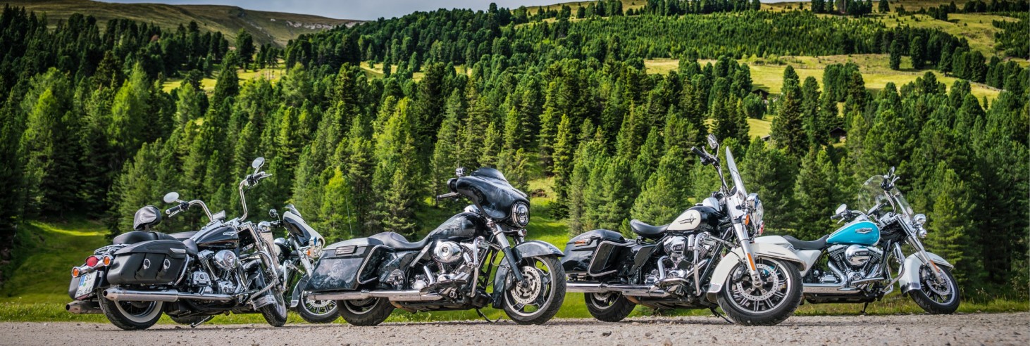 Four motorcycles in different colors are parked on a gravel road in front of wooded hills under a cloudy sky.