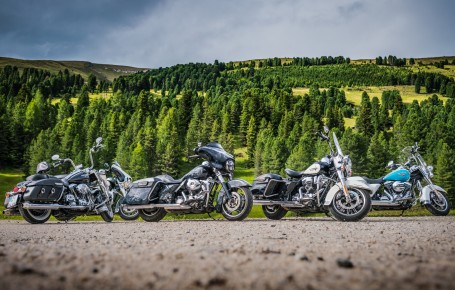Four motorcycles in different colors are parked on a gravel road in front of wooded hills under a cloudy sky.