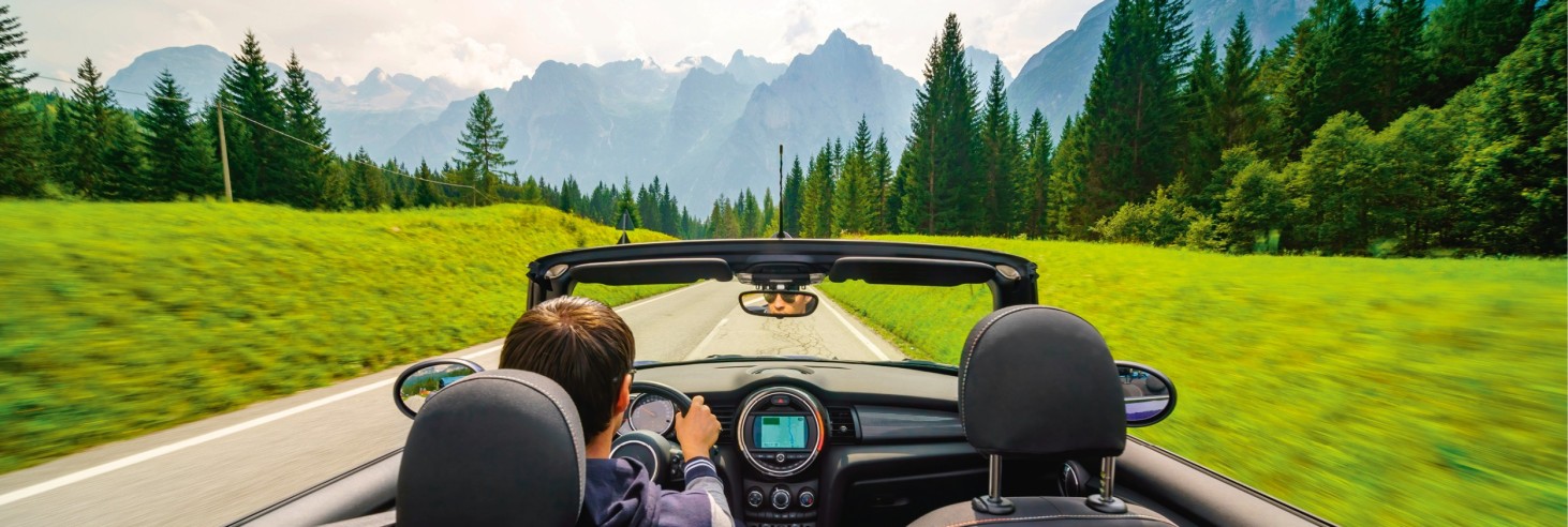 View from a convertible on a road through green meadows and coniferous forests with a mountain range in the background, driver with dark hair visible from behind.