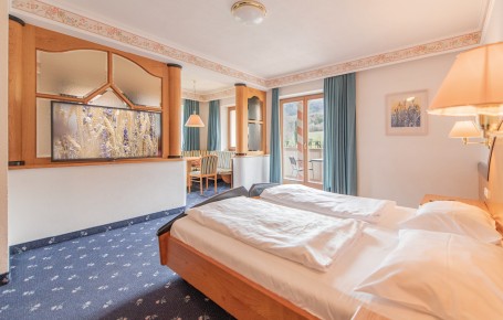 Hell eingerichtetes Hotelzimmer mit Doppelbett, zwei Nachttischen, Fernseher an der Wand, Sitzecke und Teppichboden mit Muster.